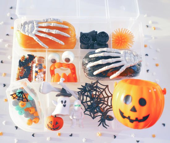 A Halloween-themed sensory play kit in a clear plastic organizer tray, featuring orange and black slime, skeleton hands, a jack-o'-lantern, spiderwebs, a ghost figure, candy corn, and small decorative accessories on a white background with scattered black and orange confetti.