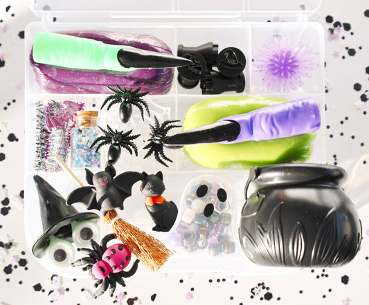 A Halloween-themed sensory play kit in a clear plastic organizer tray, featuring purple and lime green slime, black spiderwebs, plastic bats and spiders, a witch hat, a broomstick, ghost beads, and a black cauldron, all arranged on a white background with black confetti.