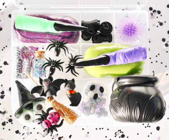 A Halloween-themed sensory play kit in a clear plastic organizer tray, featuring purple and lime green slime, black spiderwebs, plastic bats and spiders, a witch hat, a broomstick, ghost beads, and a black cauldron, all arranged on a white background with black confetti.