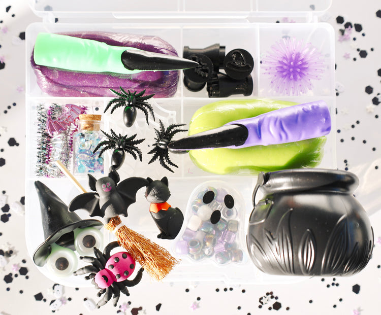 A Halloween-themed sensory play kit in a clear plastic organizer tray, featuring purple and lime green slime, black spiderwebs, plastic bats and spiders, a witch hat, a broomstick, ghost beads, and a black cauldron, all arranged on a white background with black confetti.